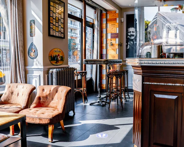 Interior view of Le Nouveau Paris with a wooden counter, high stools, and brown leather-style armchairs bathed in sunlight behind large bay windows