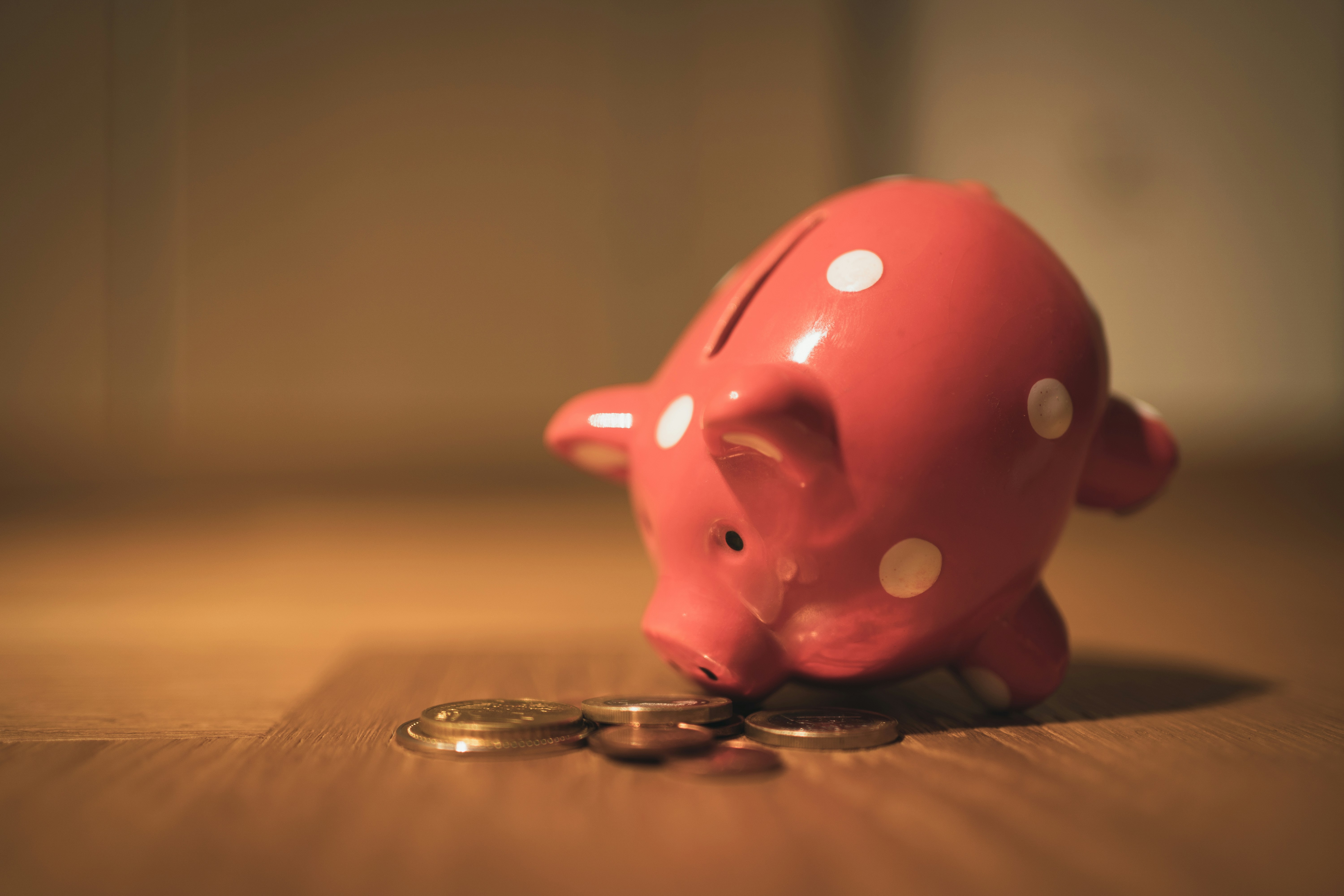 red-pink piggy bank with white polka dots tilted toward coins on a wooden surface