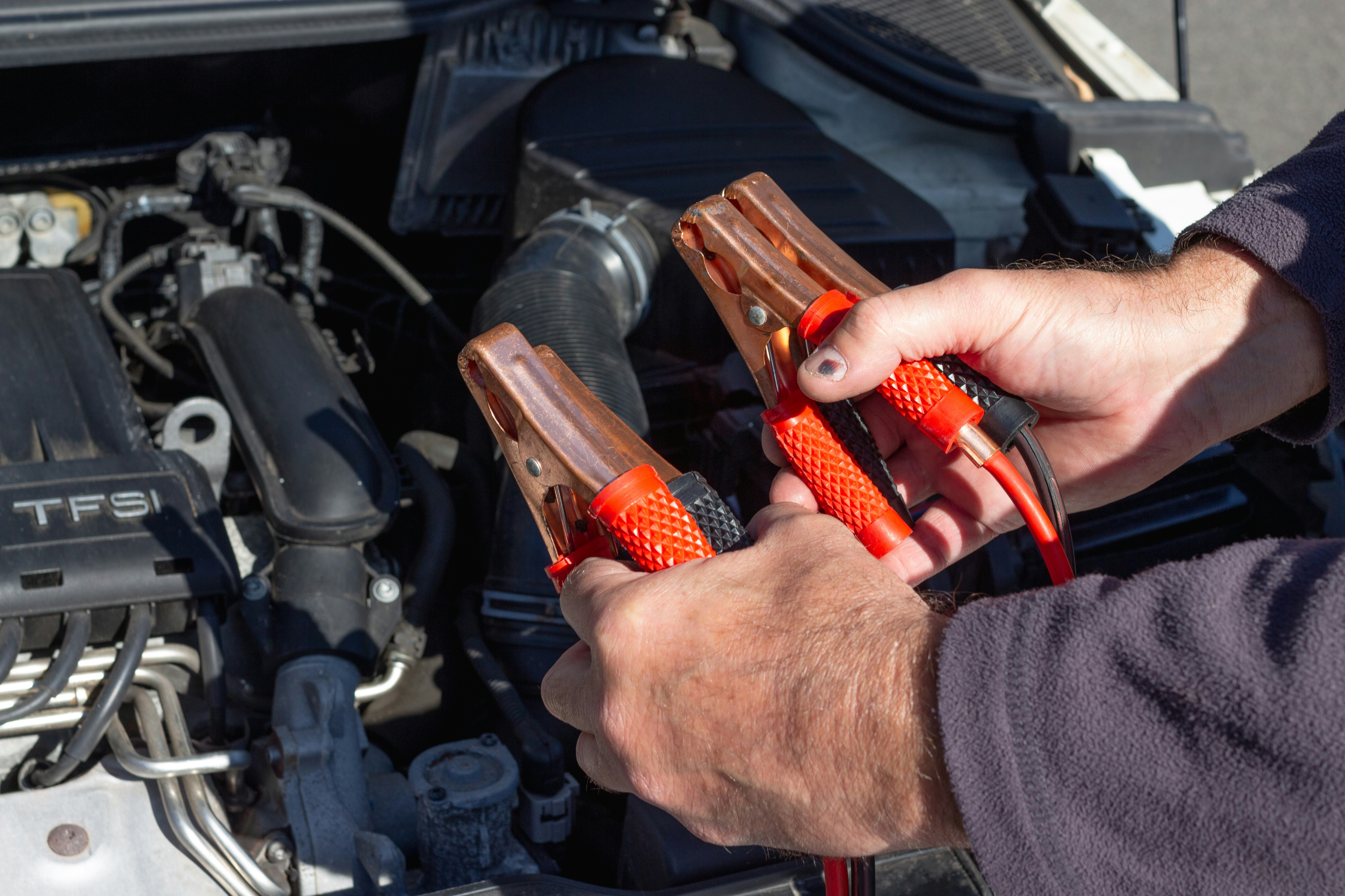 male hands holding orange-red jumper cables with open car hood in background
