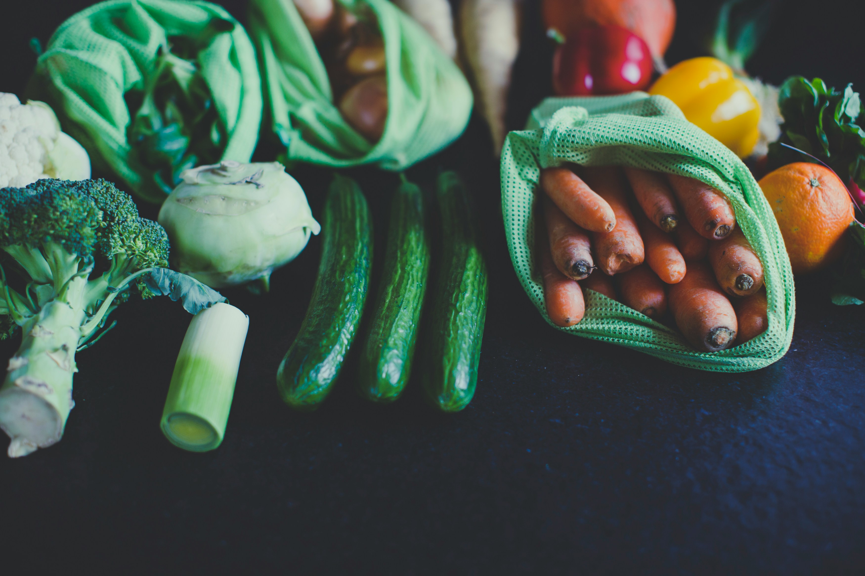 moody shot of a variety of vegetables and a few fruit on a dark blue background, including broccoli, carrots, leek, and cucumbers
