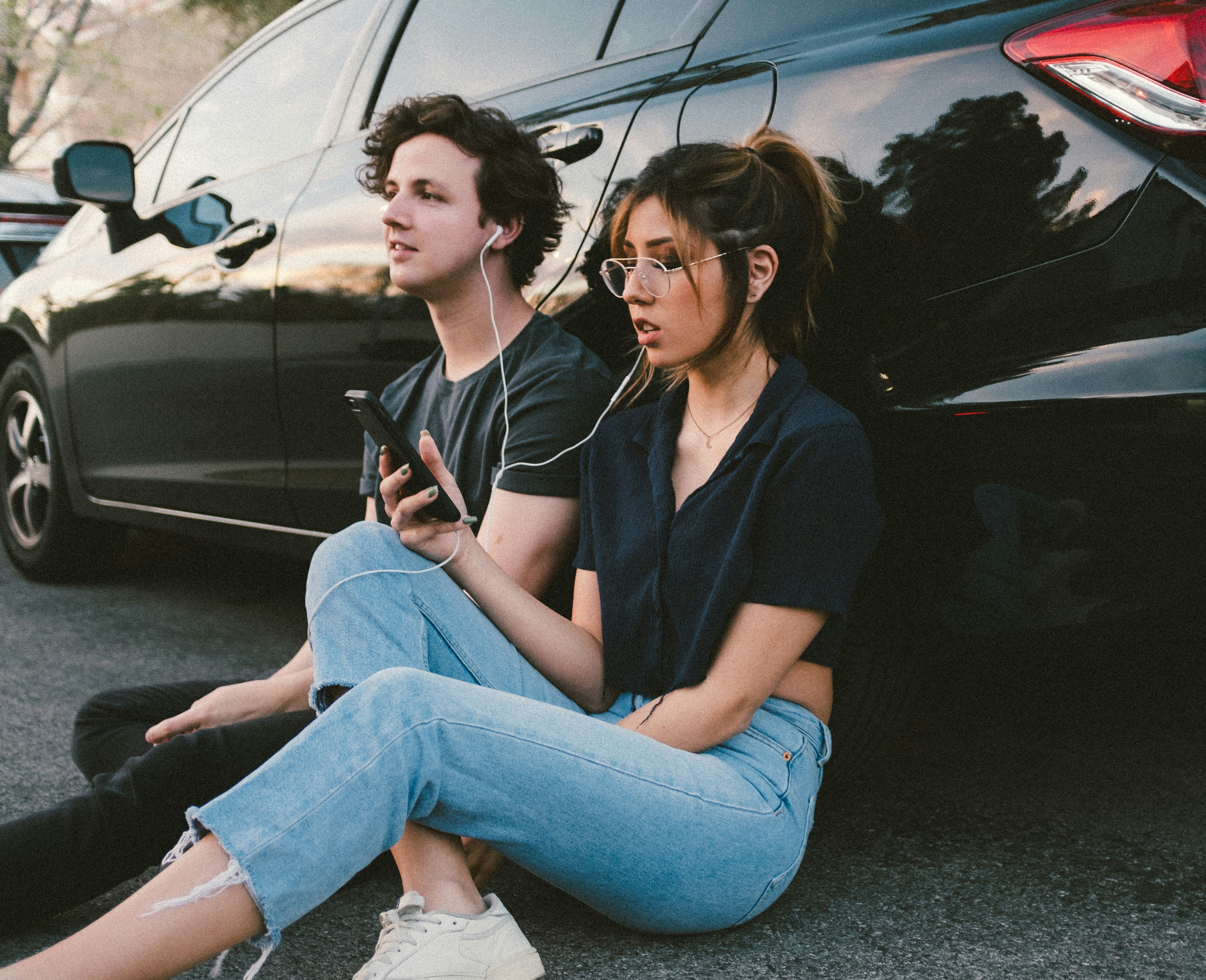 young man and woman sharing earbuds while leaning on a car