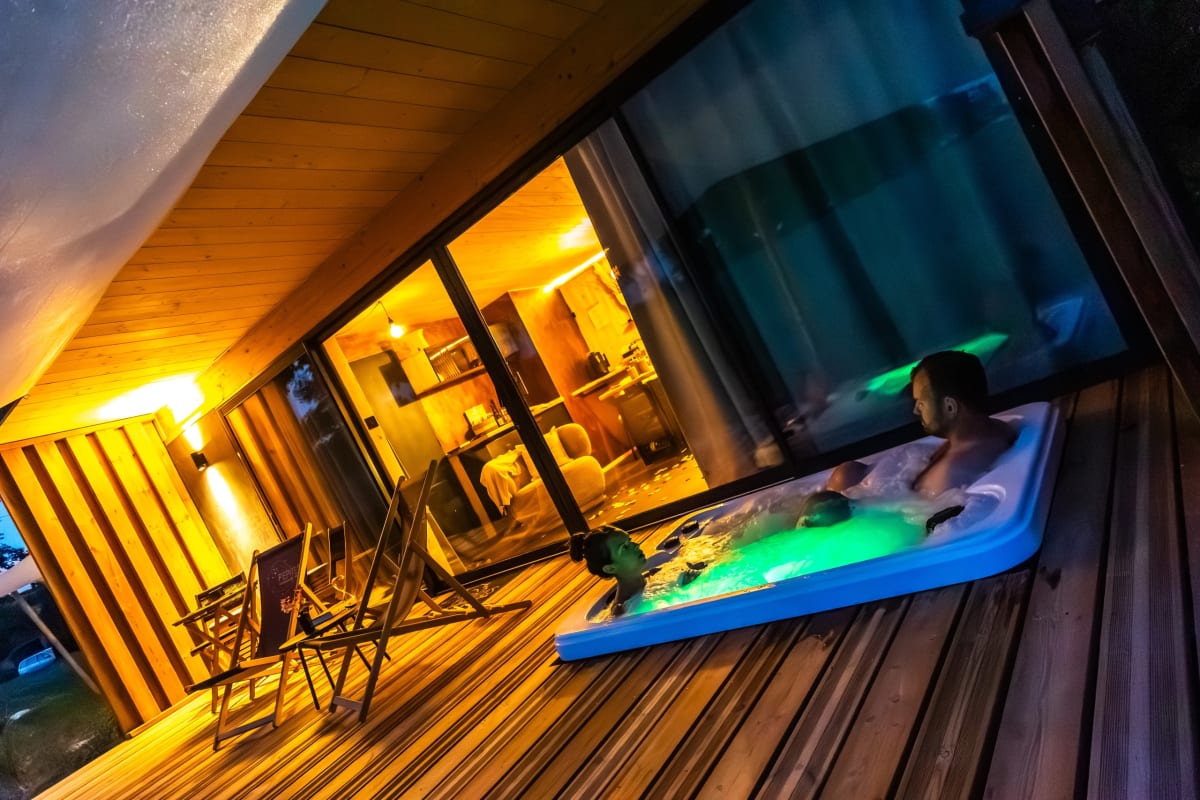Spa privatif éclairé à la nuit tombée sur la terrasse en bois d'un lodge Pépites Lodges & Spas à Prémilhat, près de Montluçon