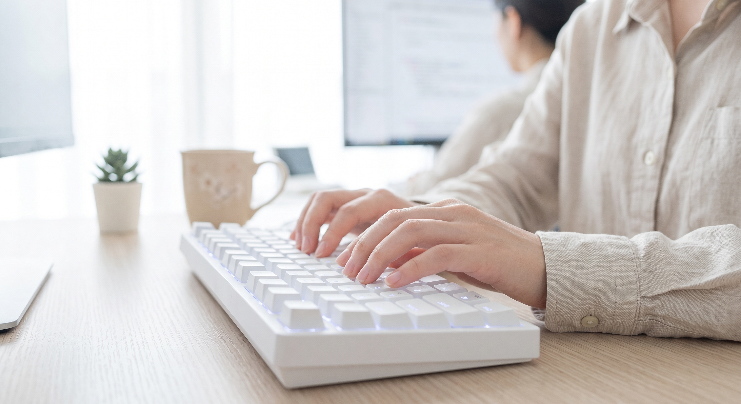 A woman typing on a white mechanical keyboard on a wooden desk.