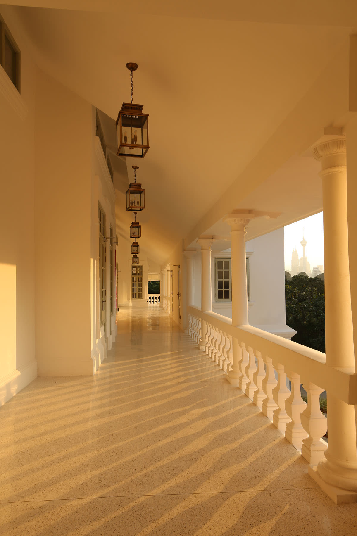 Sunlit colonnaded verandah at Seri Negara featuring white columns, hanging lanterns, and Kuala Lumpur skyline views.