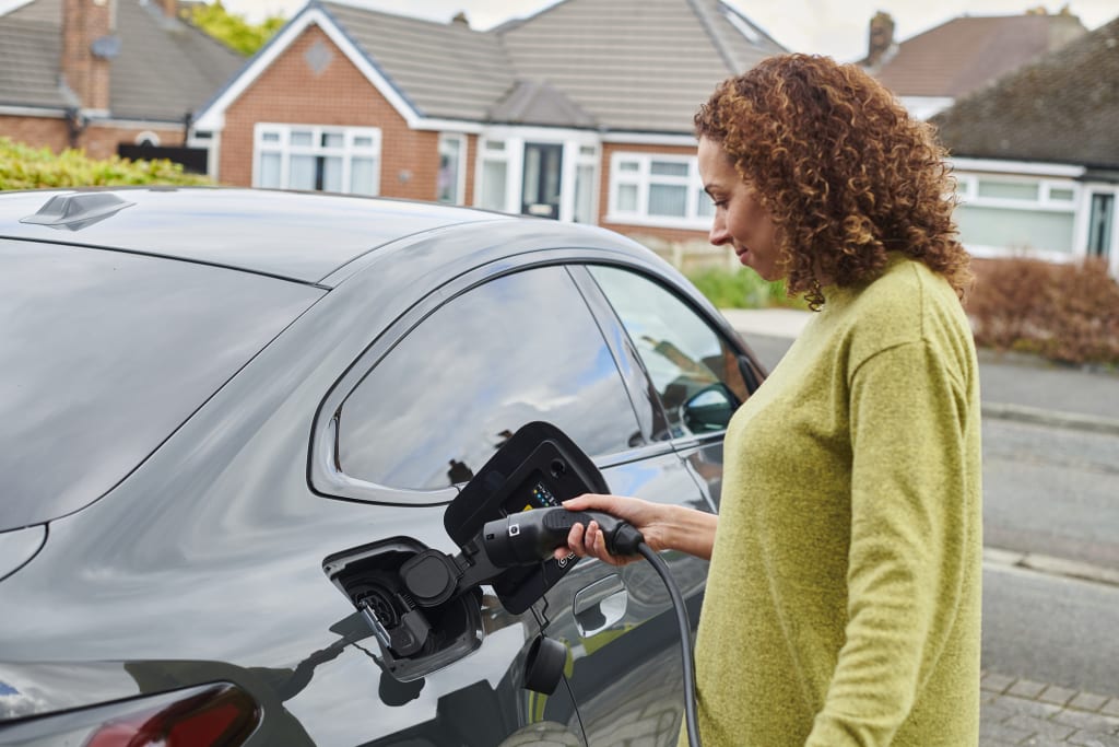 Woman with curly hair and wearing a green jumper plugging a car charger into an electric car parked in her driveway.
