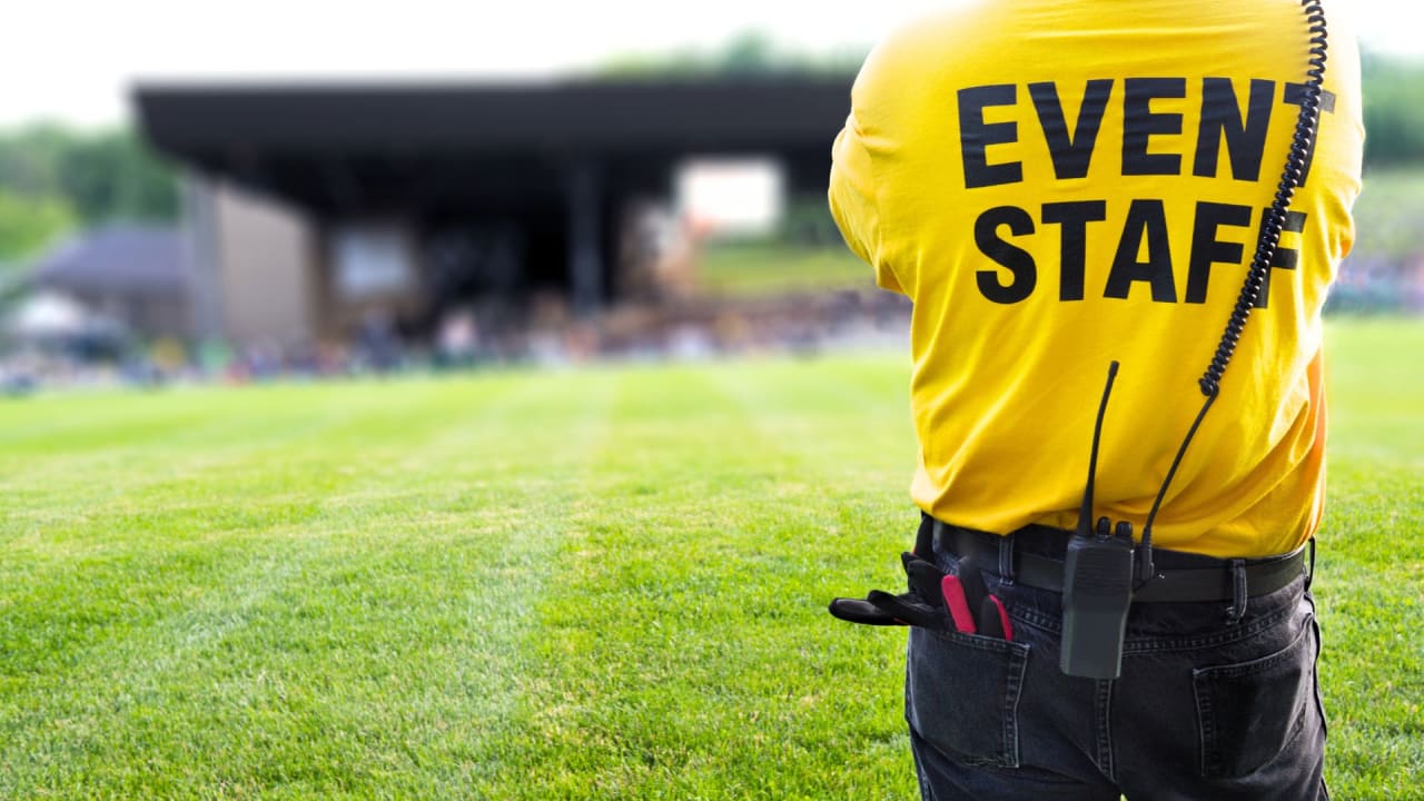 A person wearing a yellow "event staff" shirt and a Walkie-Talkie stands in front of an outdoor stage at a music festival. 