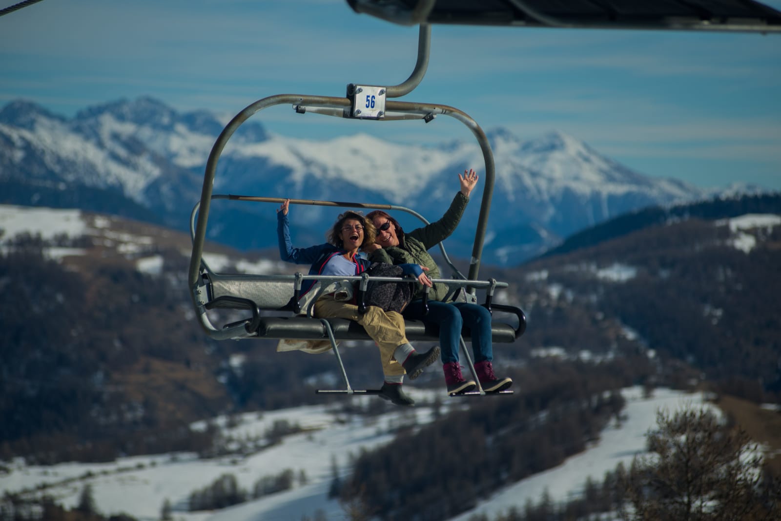 Two friends laughing on a chairlift above snowy mountains