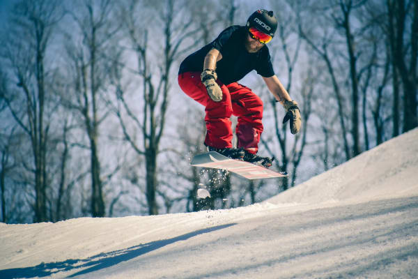 Dynamic snowboarder catching air on a snowy slope surrounded by bare trees. Capturing the thrill of winter sports.