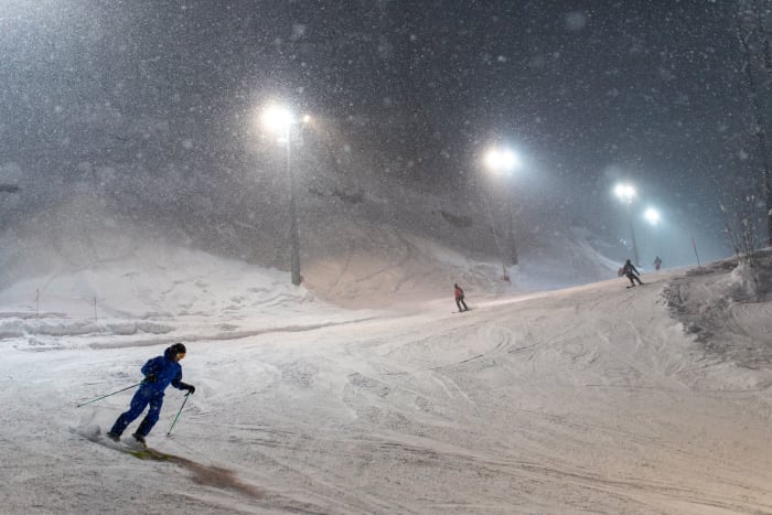 A vivid scene of skiers enjoying a snowy night on a well-lit, snow-covered mountain slope.