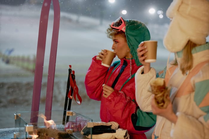 Two women in winter attire enjoying hot beverages by the ski slopes at night.