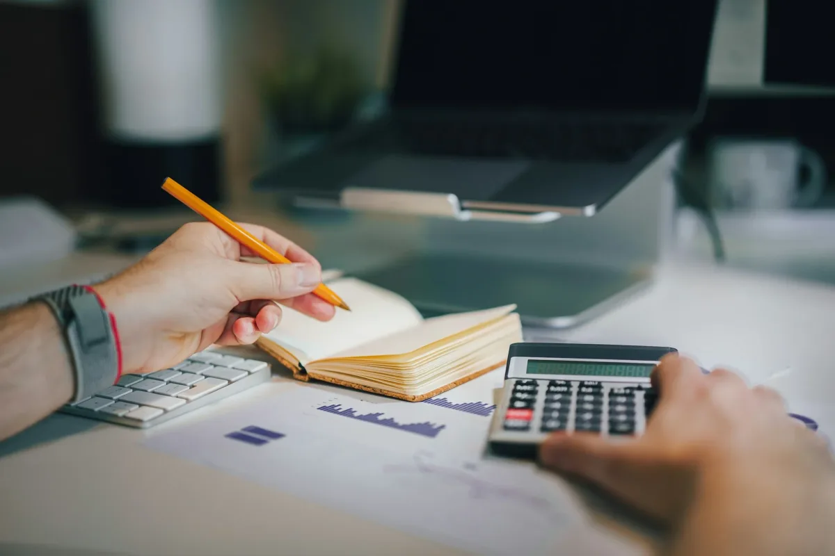 Mains calculant un budget de projet avec une calculatrice, un carnet de notes et un graphique de coûts sur un bureau