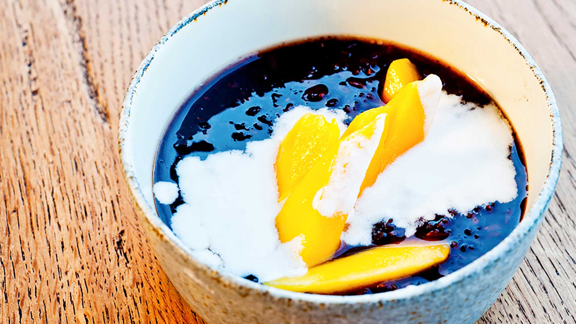 A bowl of black sticky rice pudding topped with fresh mango slices and coconut milk on a wooden table.