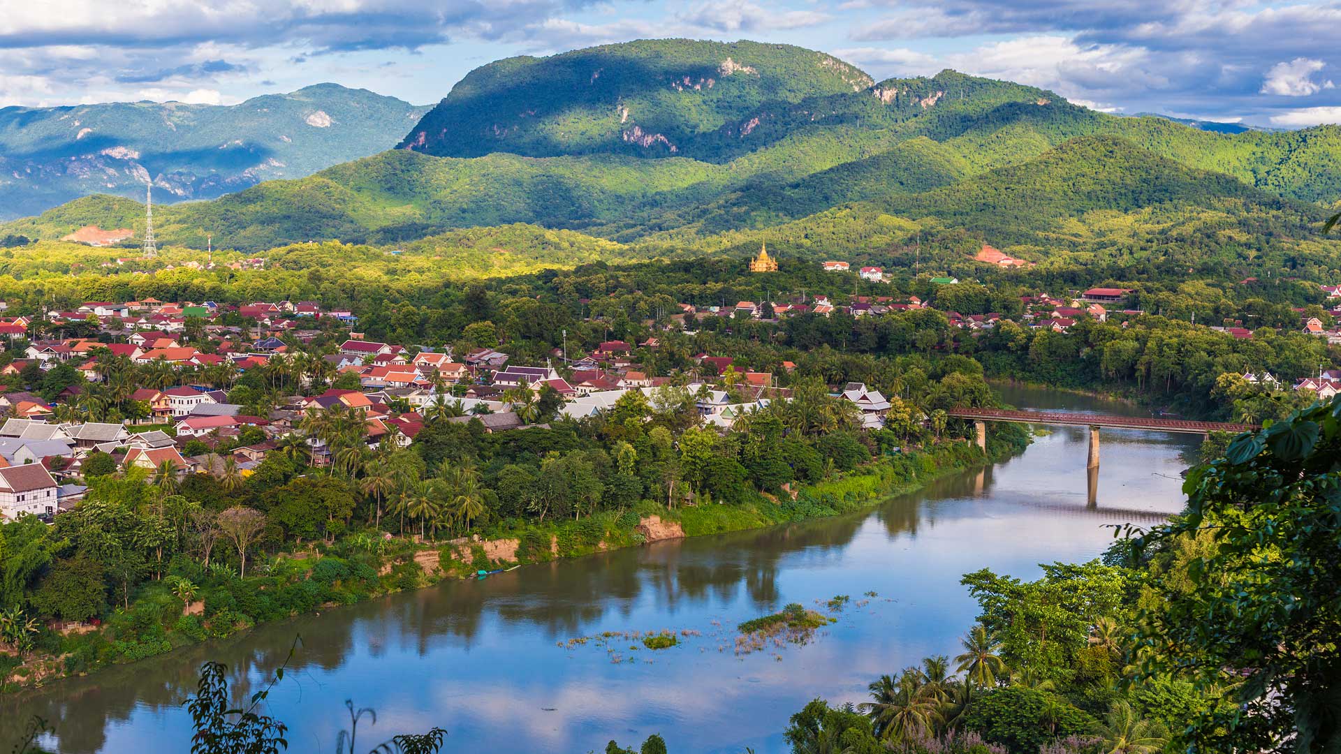 Panoramic view of a wide river flowing past a lush green town nested under rolling mountain peaks.