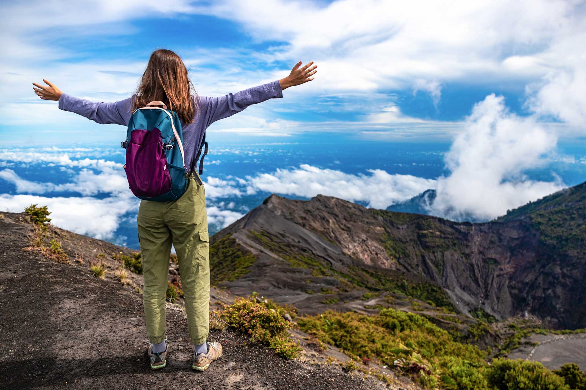A hiker stands with arms outstretched at the summit of a volcano, looking out over a lush landscape.