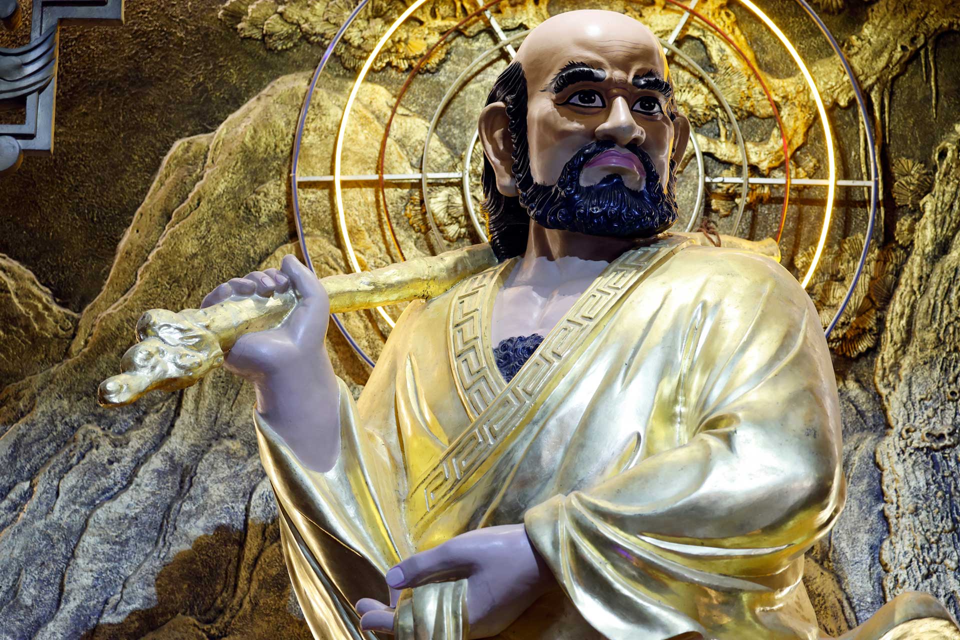 Close-up of a colorful Buddhist statue in a golden robe, holding a staff, set against a textured gold background.