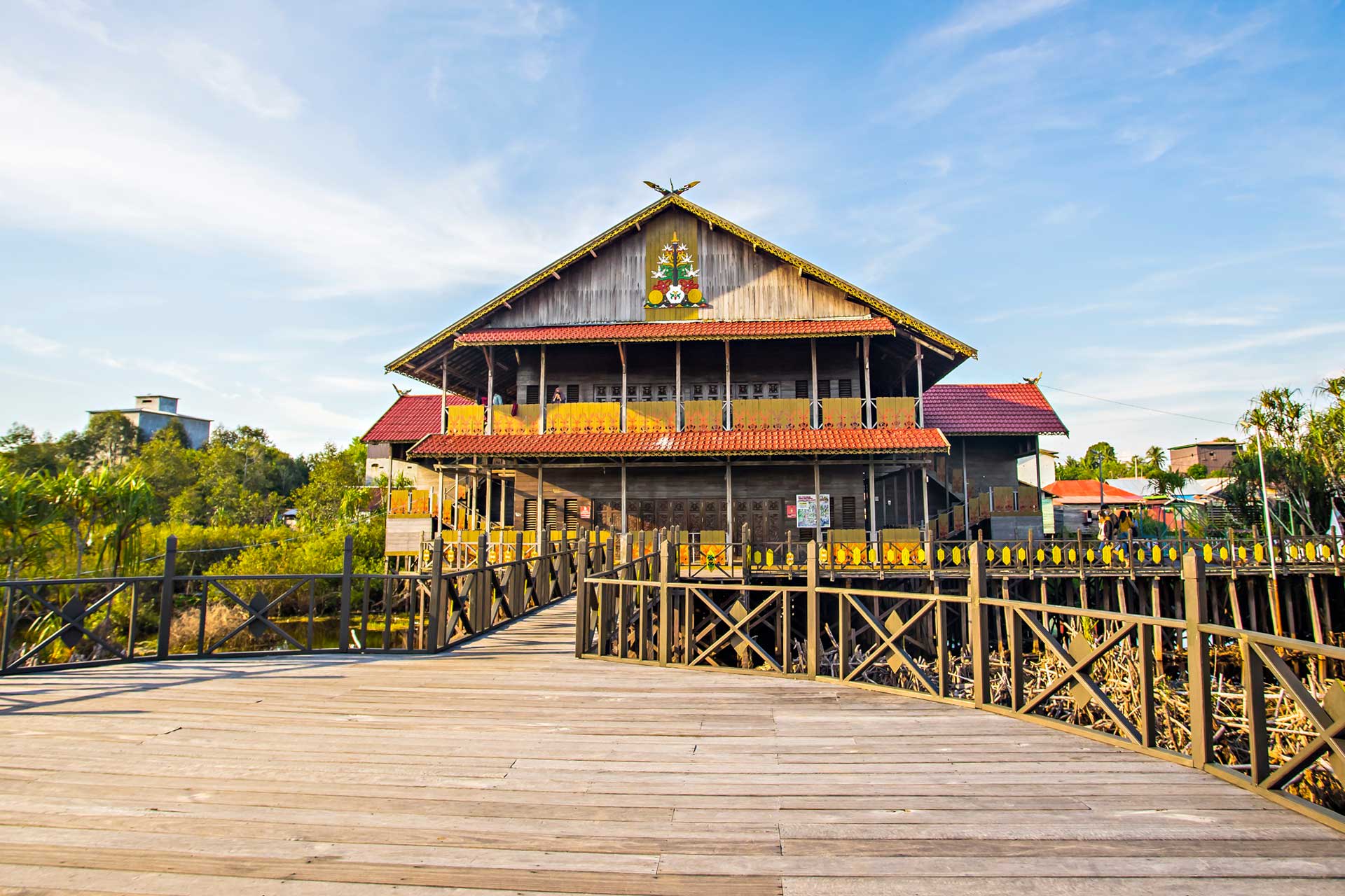 A traditional Dayak large wooden stilt house in Borneo with an ornate roof emblem, connected by a wooden boardwalk.