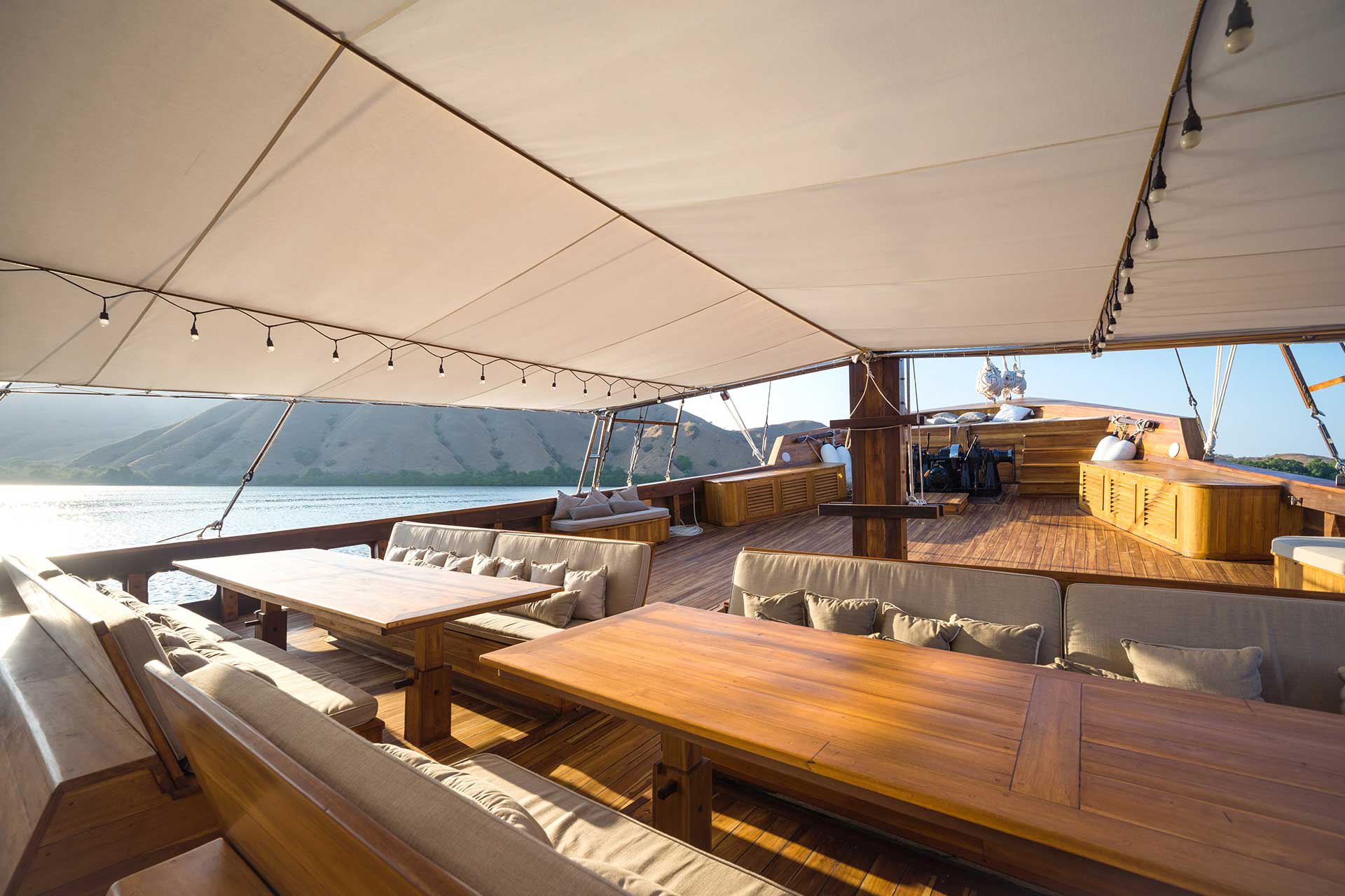 An open-air wooden sundeck on a phinisi boat featuring dining tables and cushioned seating under a large canopy.