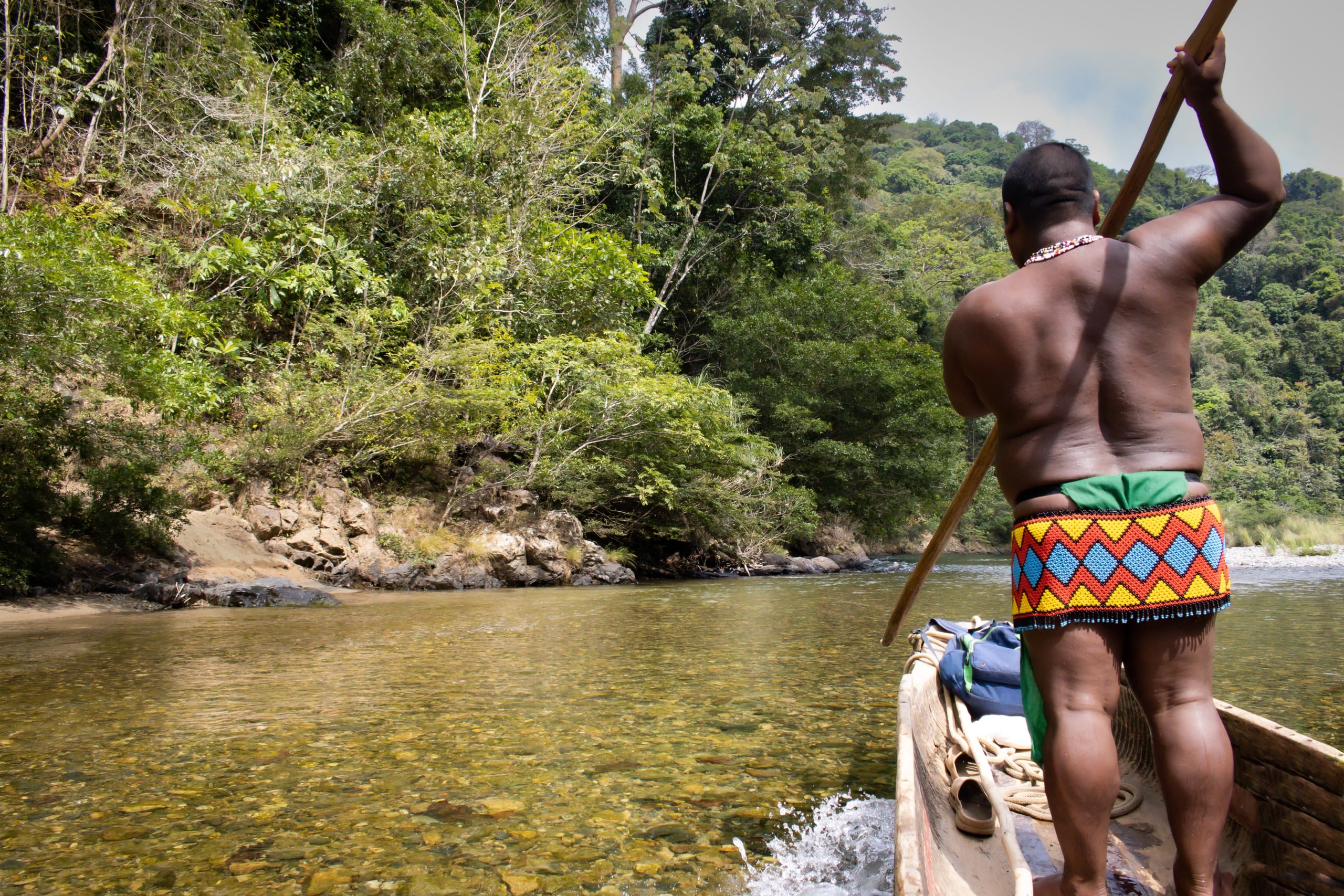 An Emberá man in traditional beaded clothing navigates a wooden canoe down a clear river in a lush jungle.