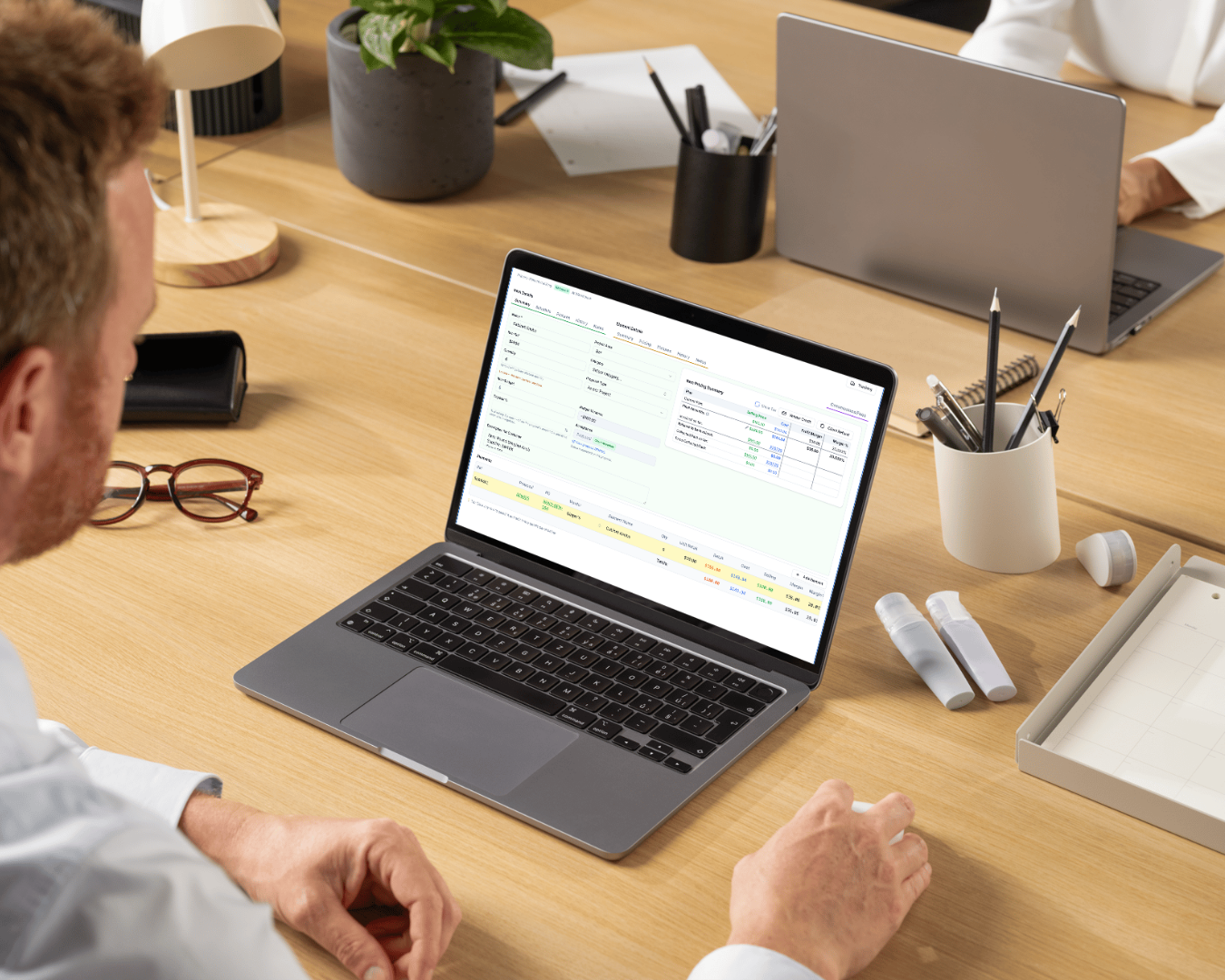 A person at a desk reviewing DesignDrive on a laptop. The screen shows an item detail view: item fields and project area on the left, and an item pricing summary panel on the right breaking out selling price, cost, profit margin and margin percentage. A colleague works at a second laptop across the desk.