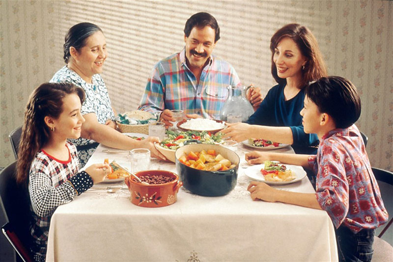A family of five enjoying a meal together around a table filled with fresh salads and hearty dishes; the atmosphere feels warm and full of togetherness
