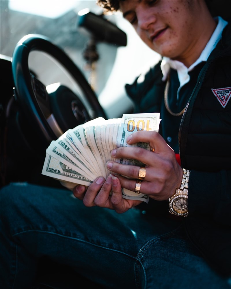 A man in a car, wearing gold rings and a flashy watch, holds a large stack of dollar bills with both hands
