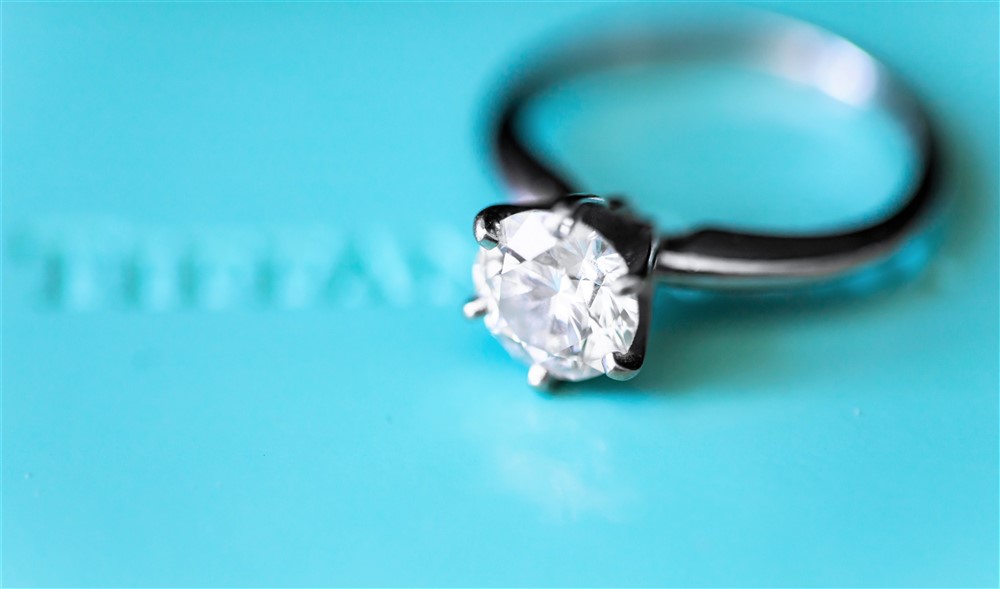 A round cut diamond engagement ring in a platinum setting, elegantly displayed against a light teal blue background