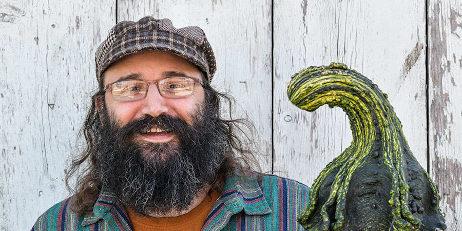 Bevin Cohen holding up a crookneck gourd in front of a weathered white wall