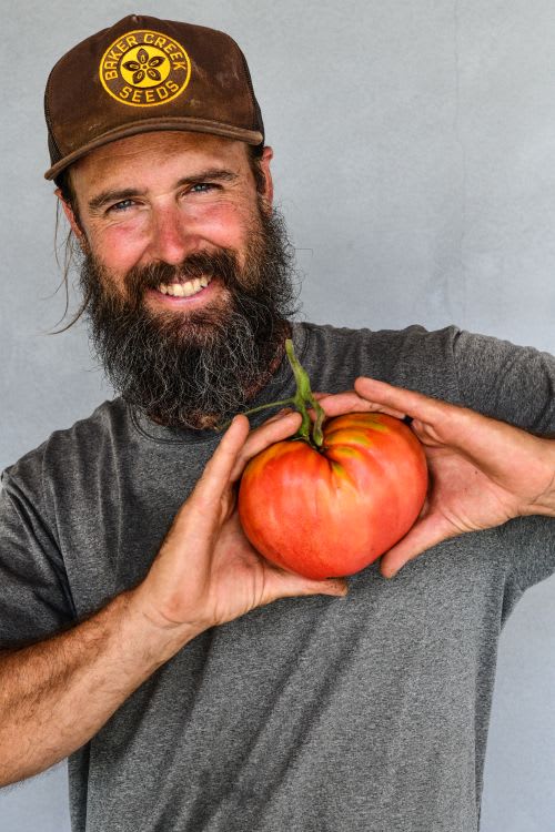 Smiling man with beard and brown cap holds a tomato