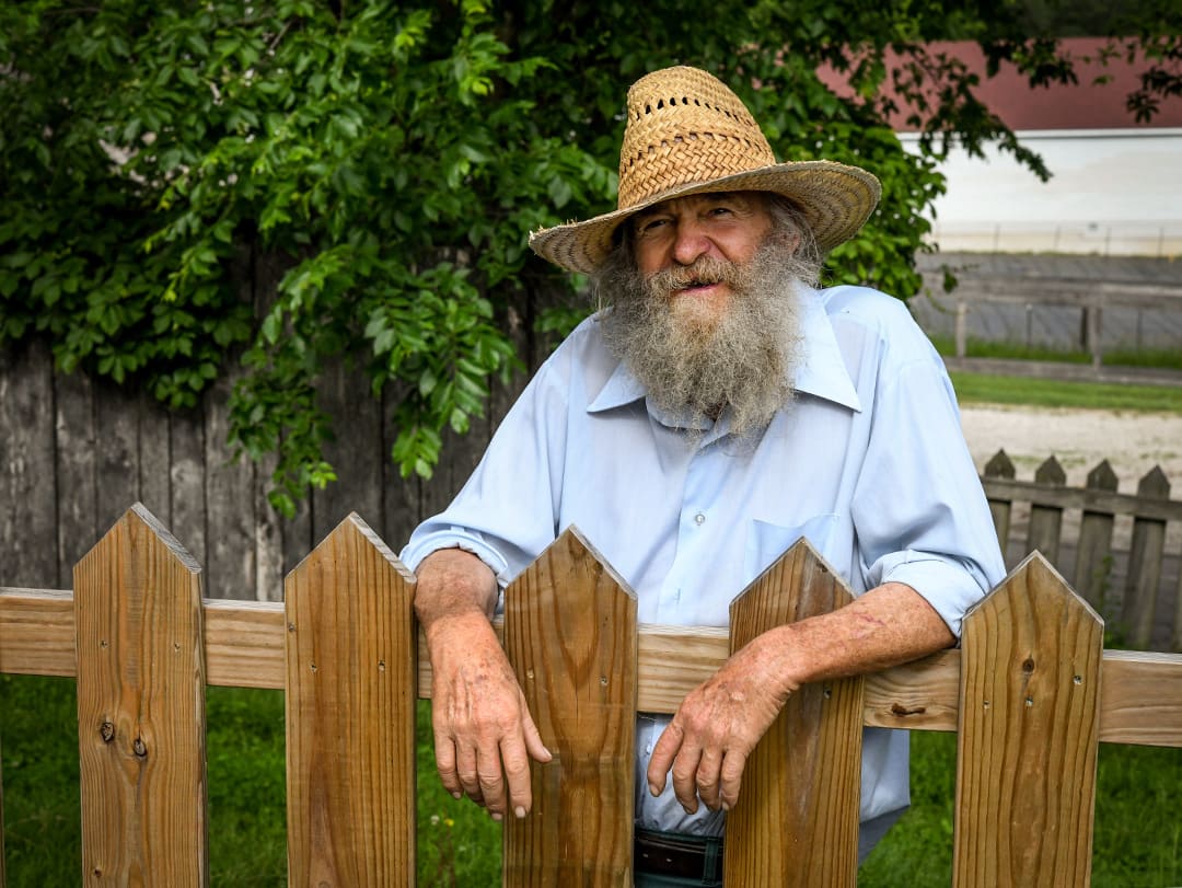 Dave, a grey-bearded man wearing a pale blue shirt and straw hat, leans on a wooden fence