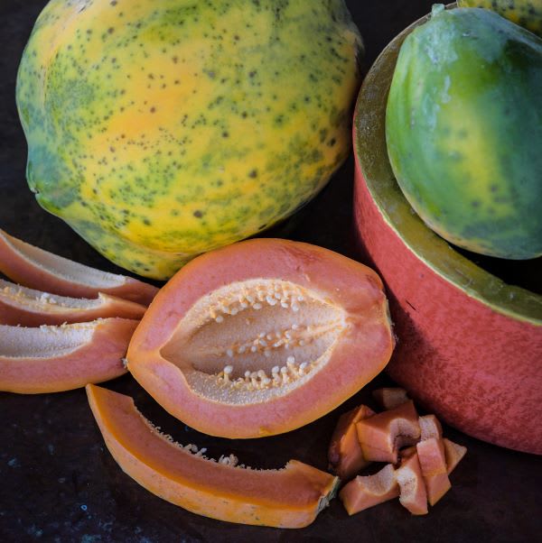several papayas are shown in close-up shot that includes slices, whole fruits and a bright orange halved fruit with pale seeds inside and some small chunks of fruit all on a dark background