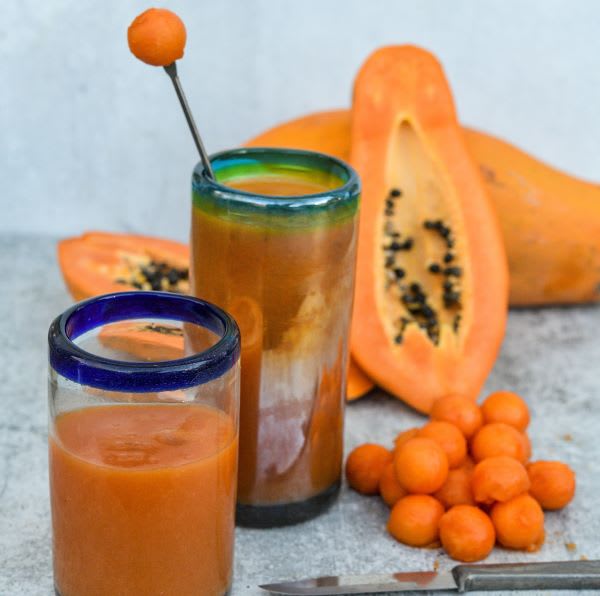 Two heavy glasses of papaya juice, one at center of image with a green rim and one in front of it with a blue rim, in front of a sliced-open ripe papaya fruit and a small stack of ripe papaya pieces, on a pale blue-grey background
