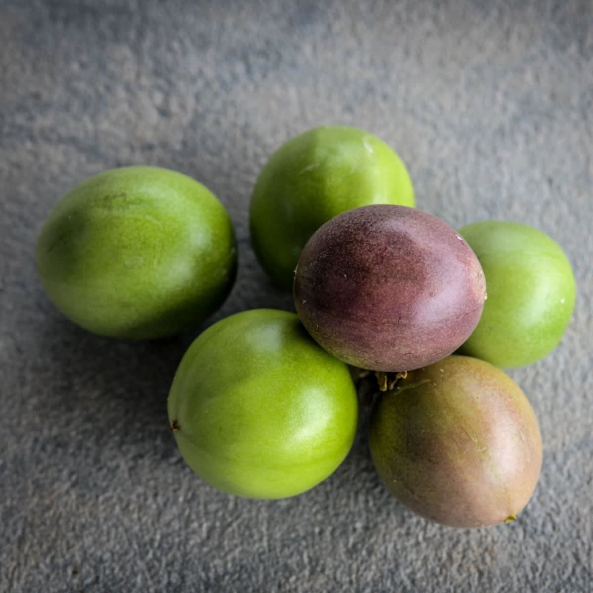 Five small round passion fruits in varying shades of green and brownish-green with a darker reddish fruit on top on a medium grey background.