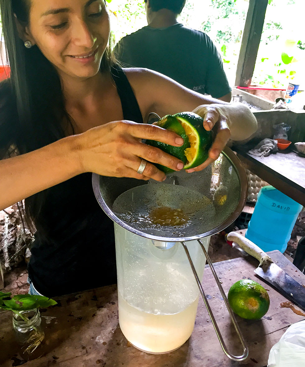 image shows a smiling woman with straight dark hair squeezing a citrus fruit into a colander in an outdoor kitchen
