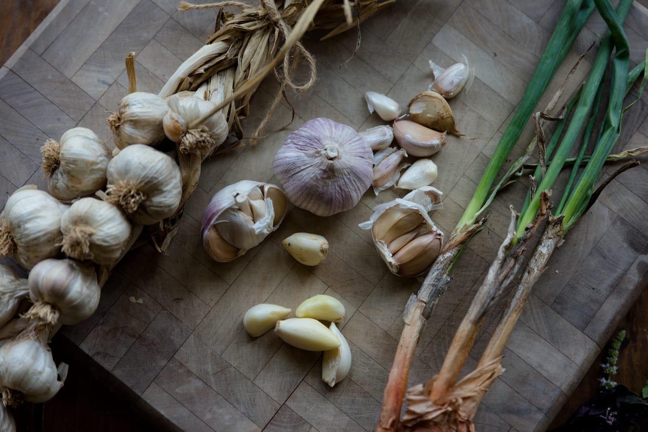 a garlic braid at left, diagonally across a wooden cutting board. Green stemmed bulbs diagonally at right; in center there are several peeled cloves, and a while garlic head and a broken head of garlic.