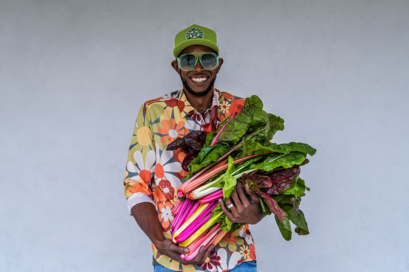 Smiling, bearded black man in flowered shirt and Baker Creek hat wearing retro-style sunglasses holds several bunches of Silverbeet swiss chard, with dark green leaves and stems of yellow, pale green, white, and several shades of pink