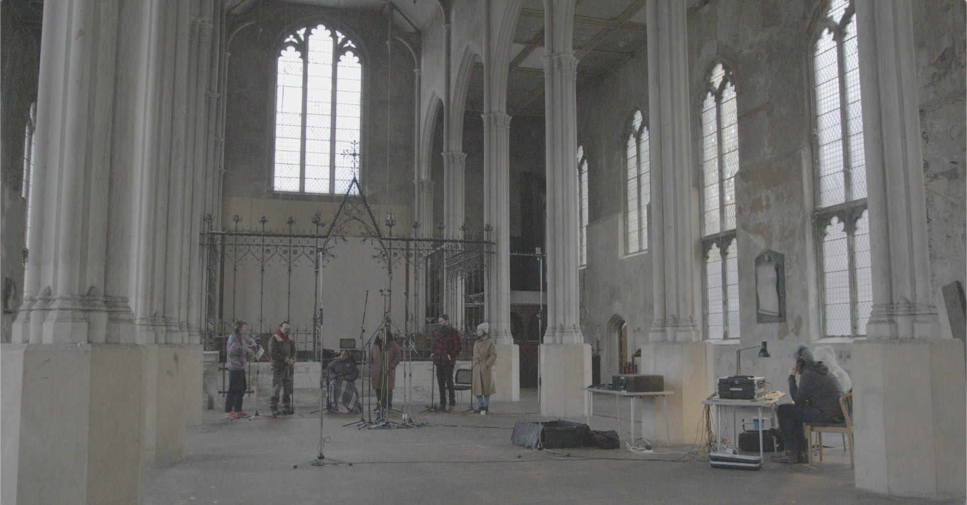 6 singers stand in a line at the end of an empty church with tall cream stone columns. Two technicians record to the right.