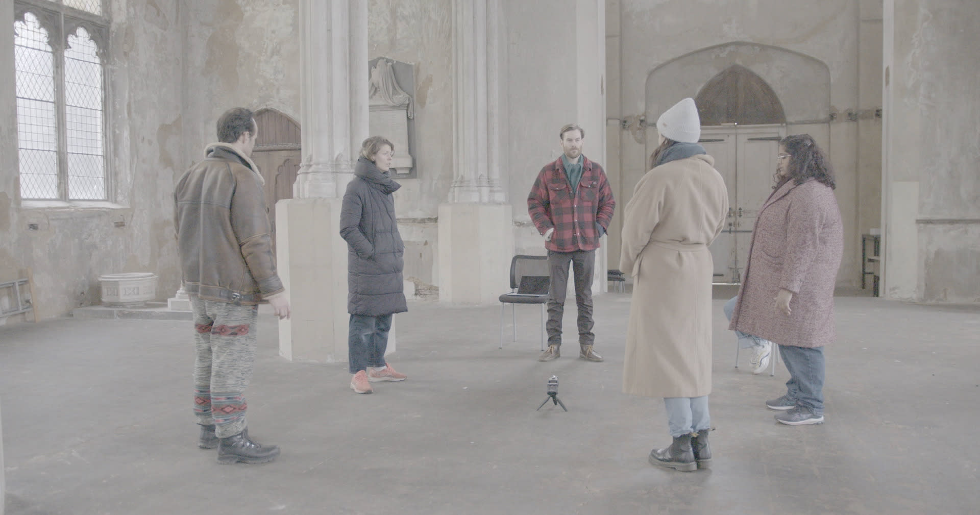 6 people stand in a circle facing each other in a large empty stone church. Some have their eyes closed. The colours are muted.