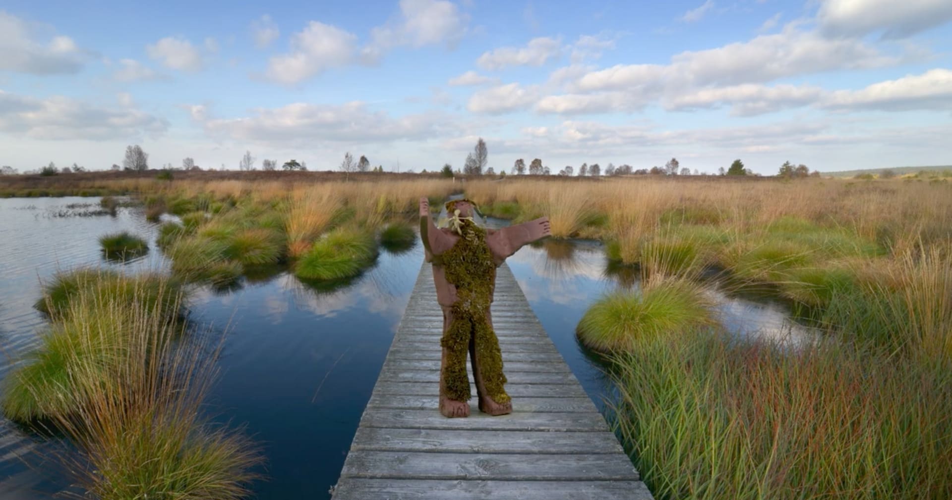 A clay figure representing a Peat-person standing on a bridge, surrounded by healthy peatlands