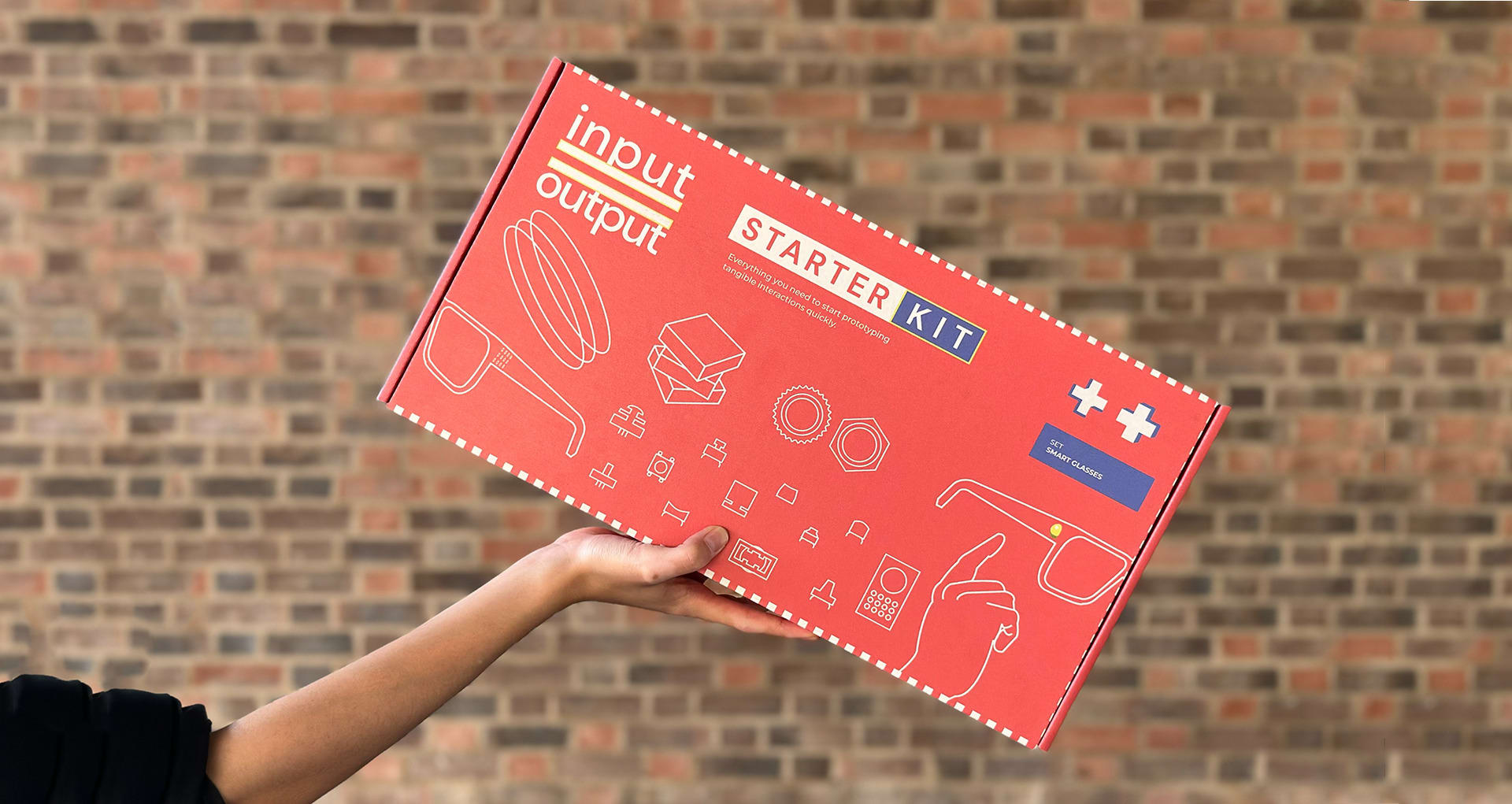 A box with "Input output starter kit" written on it. A hand holds the box in front of a brick wall.