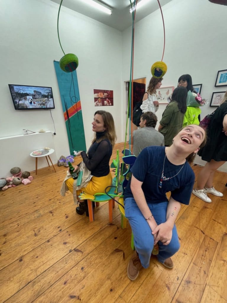 Cas sitting on an artwork, in an exhibition they curated at IMT Gallery. They are wearing blue trousers and a blue shirt.