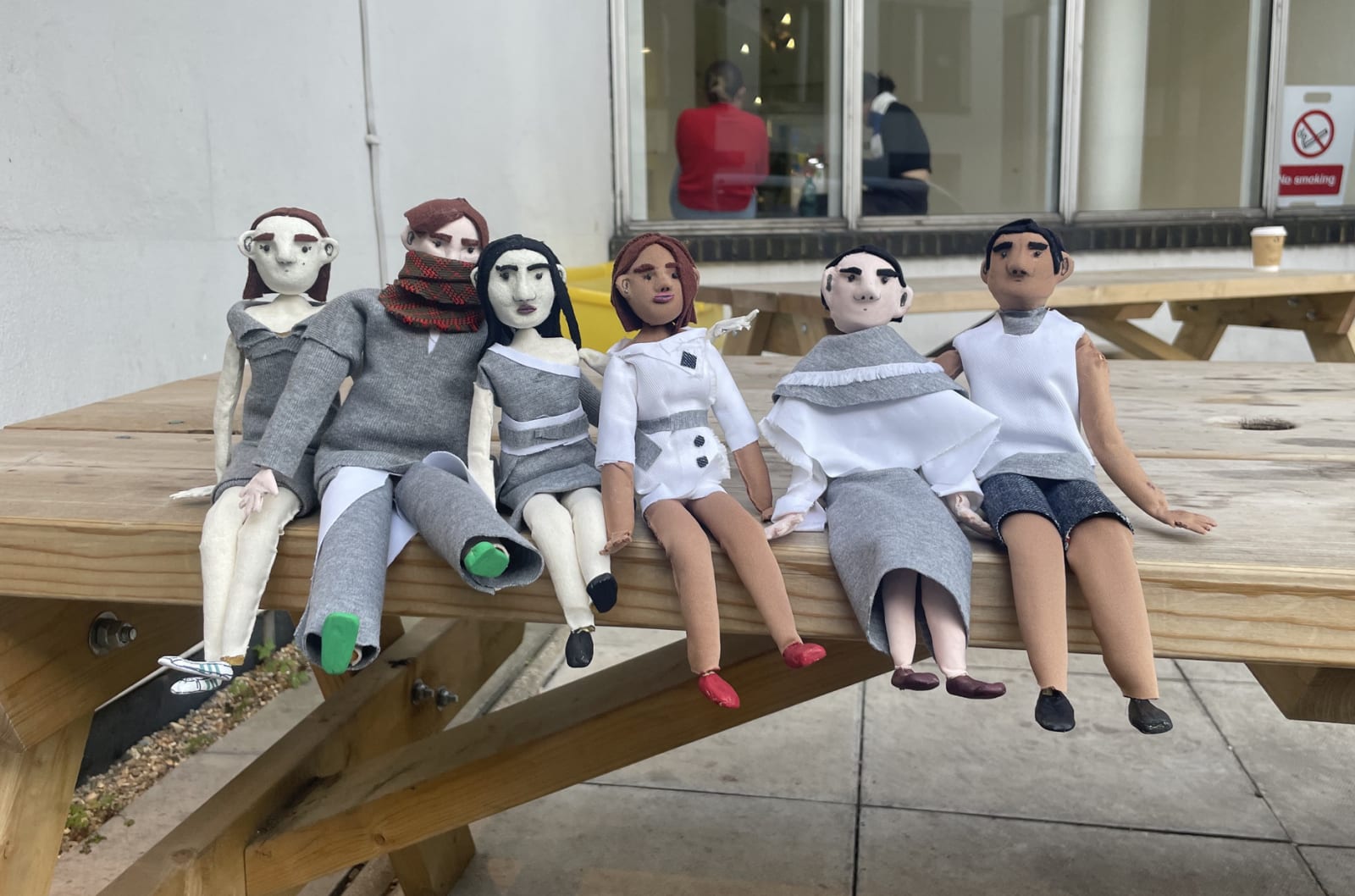 Six animation puppets, dressed in grey and white, sitting on a wooden table. Various skin colours and genders.