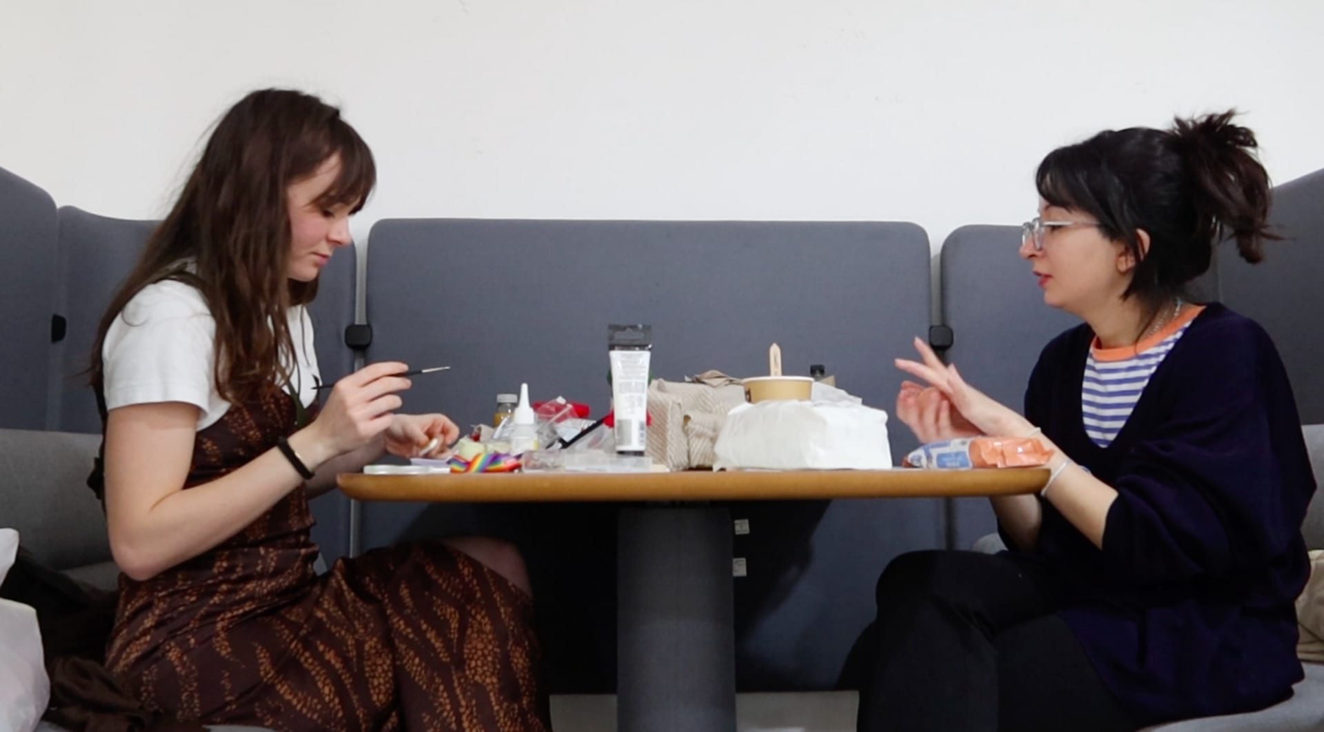 Two women sitting at a table, facing each other and painting set props 