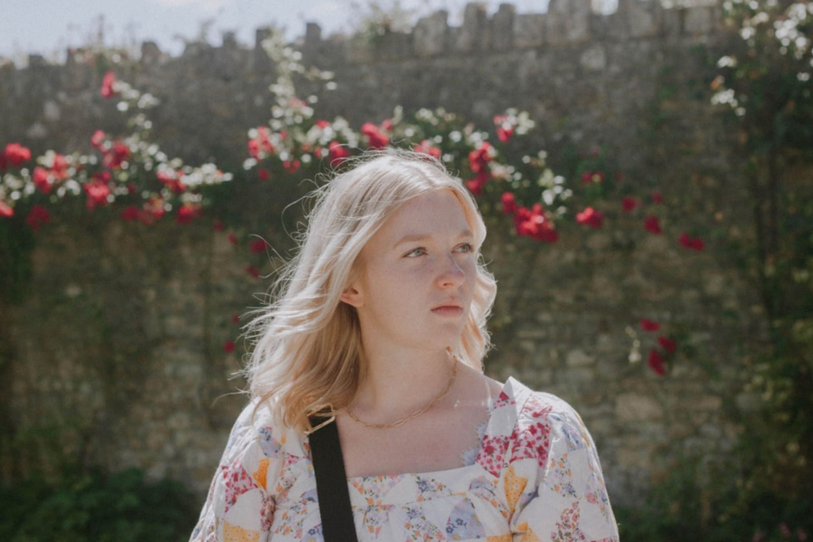 A young white woman is standing in front of a stone wall with rose climbers.