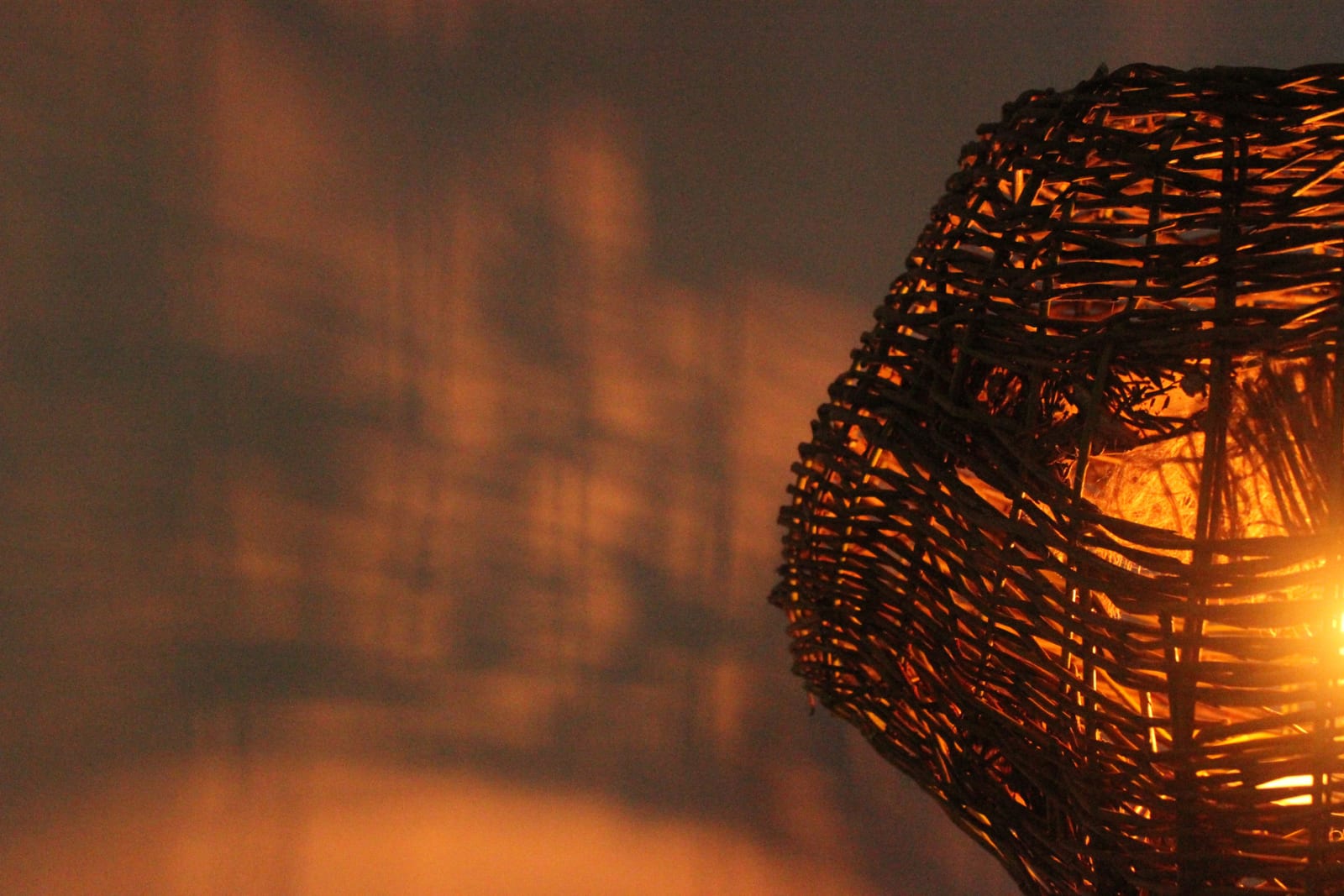 A lampshade woven using willow lit from within and casting irregular shadows on the wall behind.