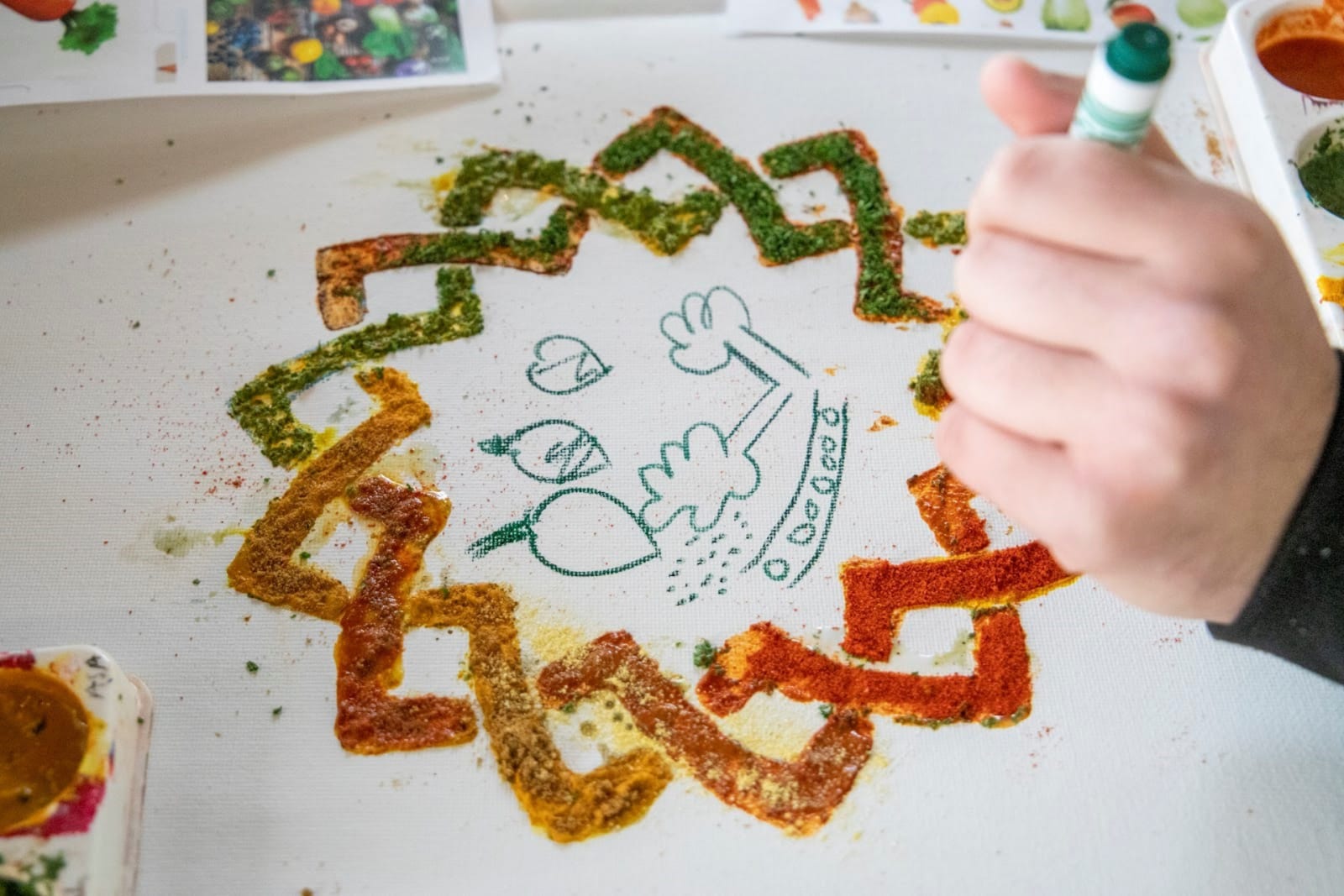 A hand clutching a green crayon is drawing on canas. The canvas showe a pattern created by sticking different spices together. 