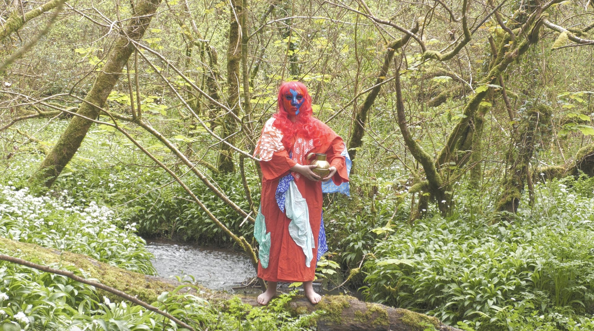 Woman stands on fallen tree branch over a stream wearing red mask, red wig and red kaftan holding a brass vase