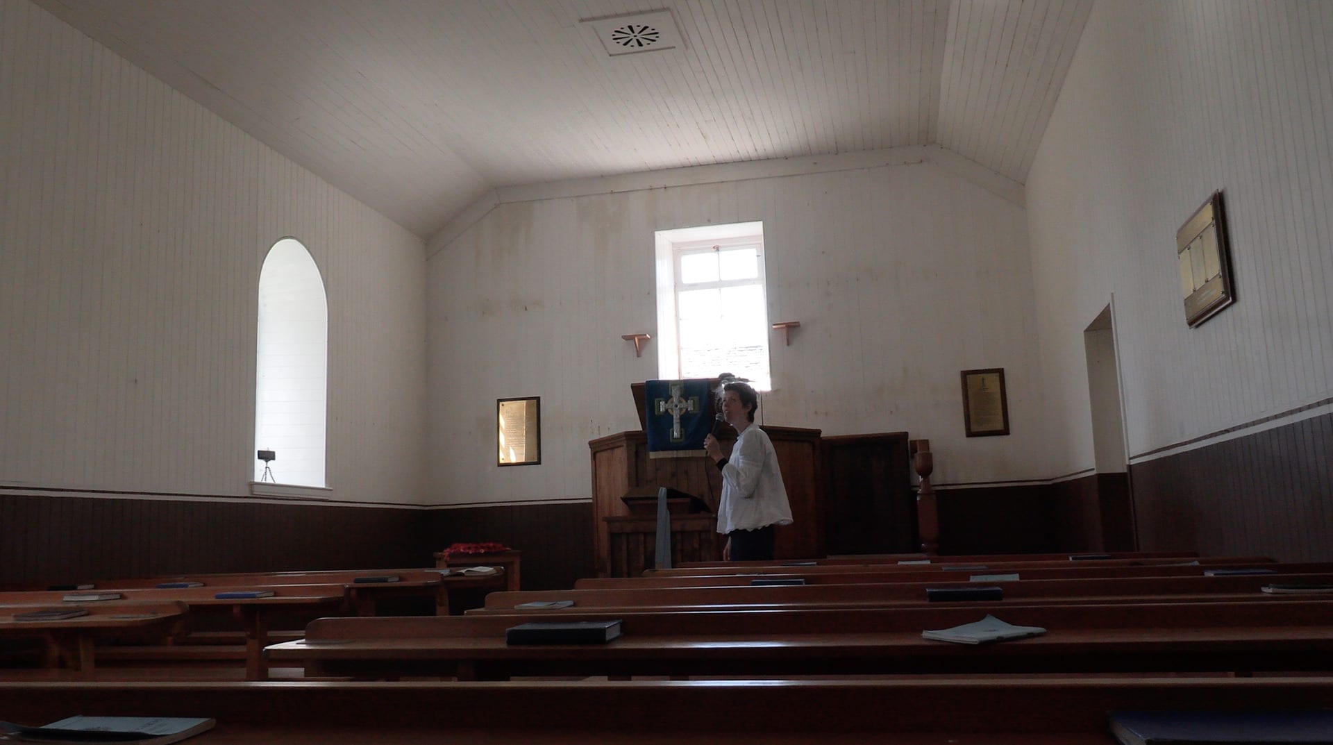 A woman sings through a microphone in a simple church. There are 2 plain windows, a white panelled ceiling and dark brown pews.