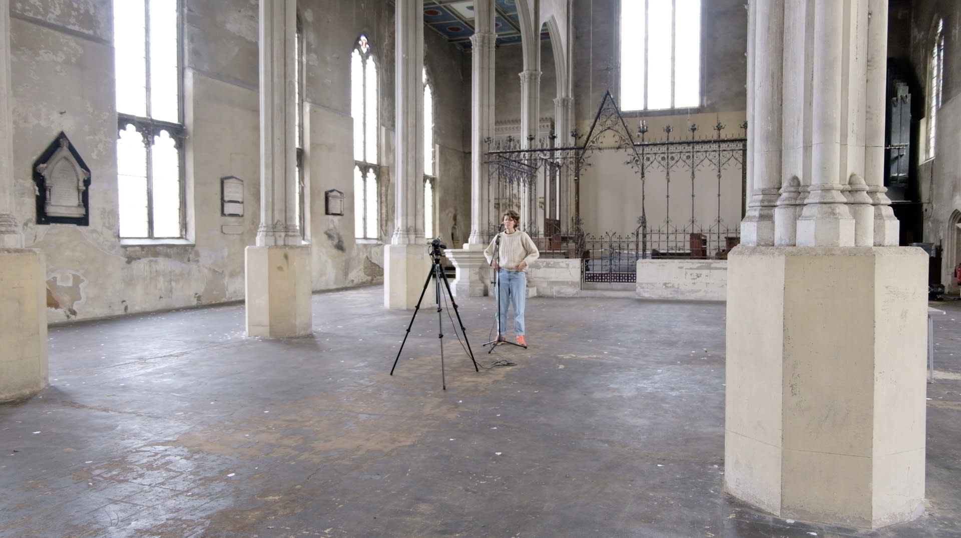 A woman stands in an old empty church singing from a microphone. The church is ornate with cream stone pillars and a wide floor.