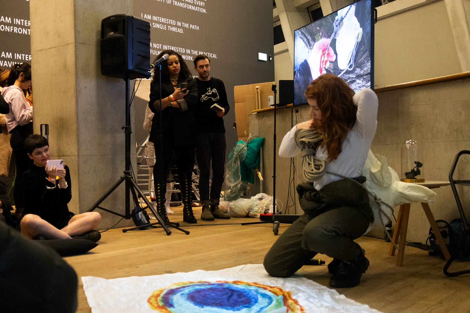 The artist performs to an audience at the Tate Modern, with a film playing on a screen in the background. They carry props.