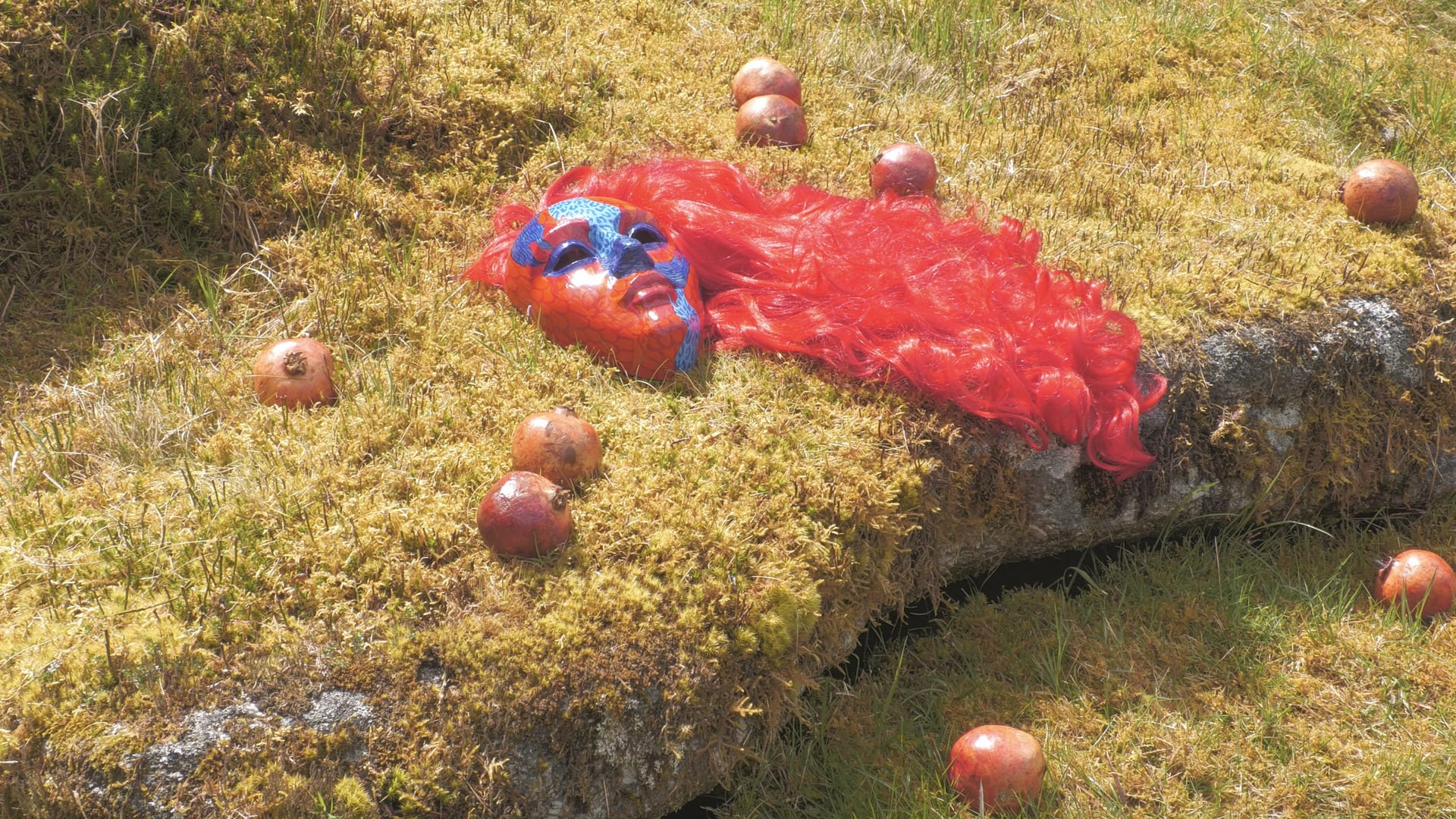 Red mask with red wig on grass surrounded by pomegranates